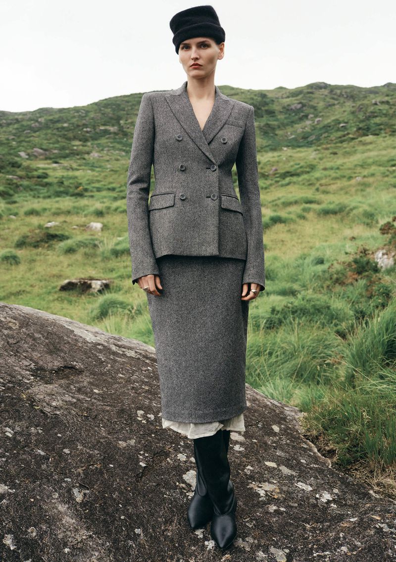 A woman in a grey tweed skirt suit with a black hat, standing on a rock, poses in a lush green field in a fashion editorial shot.