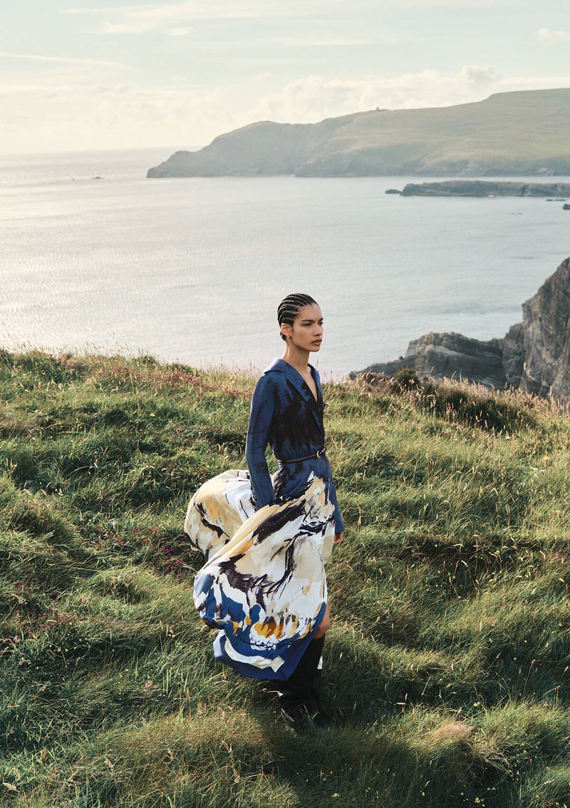 A woman in a flowing dress stands on a grassy hill overlooking the ocean, with cliffs and headlands visible in the background.