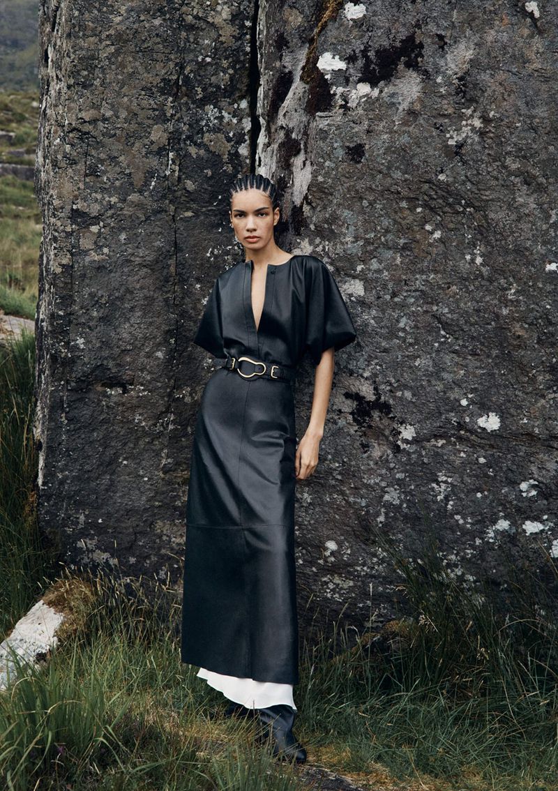 Fashion editorial shot of a woman in a black dress posing near a rock formation in an outdoor setting, showcasing modern style and nature.