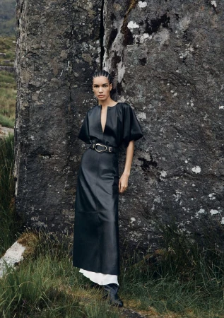 Fashion editorial shot of a woman in a black dress posing near a rock formation in an outdoor setting, showcasing modern style and nature.