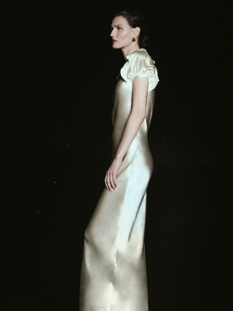 Elegant woman in satin gown stands against a dark backdrop, showcased in a fashion-style portrait with rich color and classic composition.