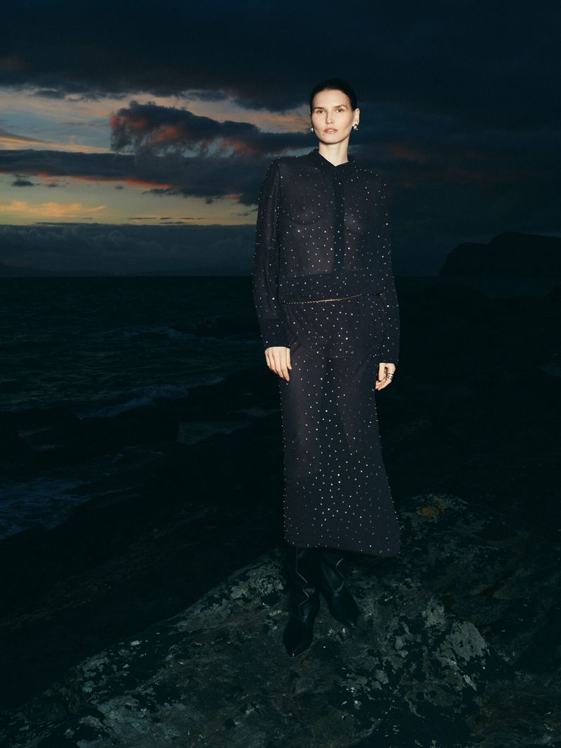 Fashion shot of a woman in a dark embellished dress standing on a rocky surface, ocean and cloudy sky in the background, dark mood.