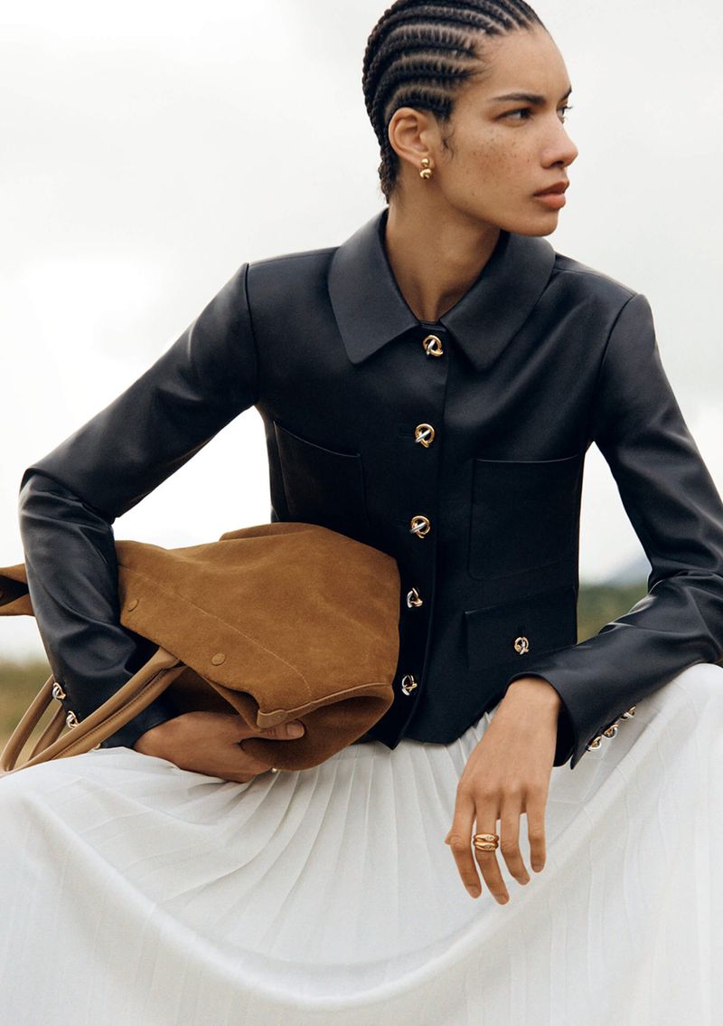 Fashion shot of woman in a black leather jacket and a white skirt holding a tan suede bag in a field, captured in natural lighting.