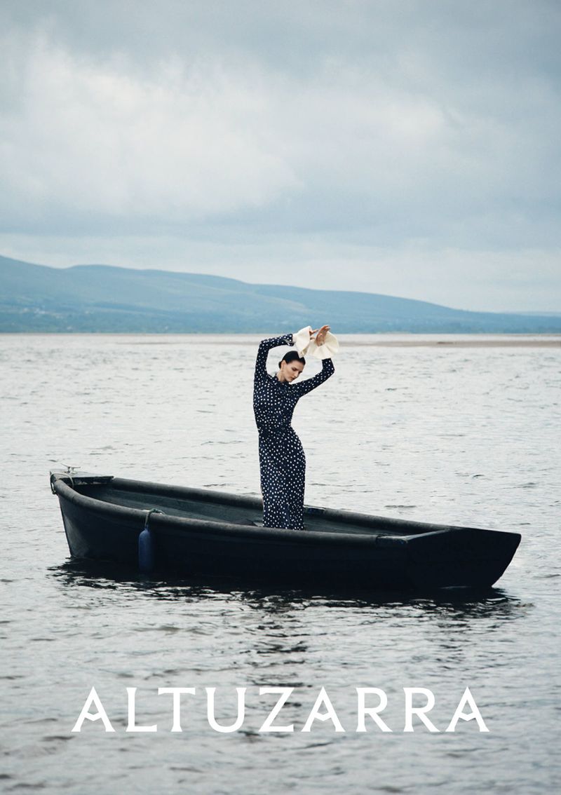 A woman in a polka dot dress stands in a black rowboat holding a hat over her head, on the water with mountains in the distance.