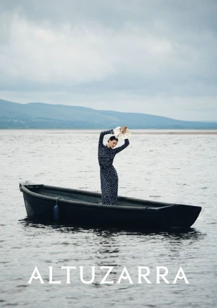 A woman in a polka dot dress stands in a black rowboat holding a hat over her head, on the water with mountains in the distance.