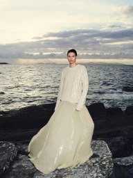 Model Katlin Aas wearing an ivory cable knit sweater and long sequin maxi skirt standing on dark rocky coast by the ocean.