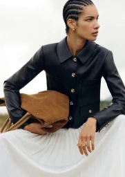Model with braided hair wearing a black leather jacket and white skirt holding a brown suede bag outdoors.