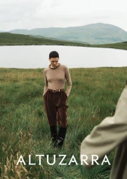 Model wearing a tan knit top and brown trousers standing in a grassy landscape by a lake for Altuzarra
