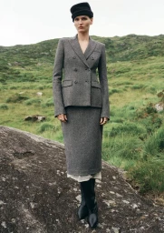 Model wearing a grey tweed double-breasted skirt suit and black beanie standing on a rock in a grassy hillside landscape.
