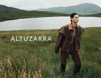 Model wearing a brown cape jacket and riding boots standing in a grassy field in front of a lake and hills.