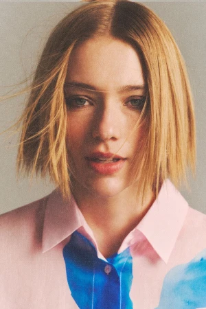 Close-up of a fair-skinned young woman with blonde bob-cut hair, wearing a pink and blue watercolor patterned collared shirt, indoors.