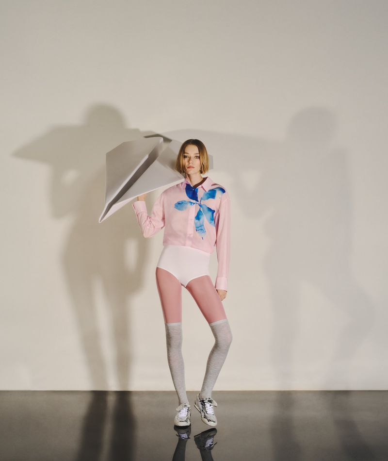 Blonde woman poses in a light pink shirt, high-waisted white shorts, and grey knee socks, holding a large, folded paper object.