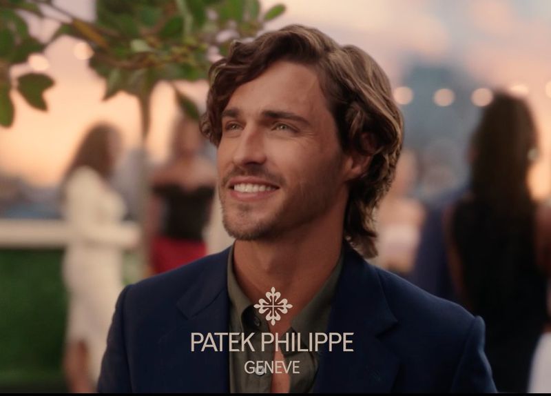 Close-up shot of a smiling man with curly brown hair wearing a navy blazer at an outdoor event during sunset hours with a crowd.