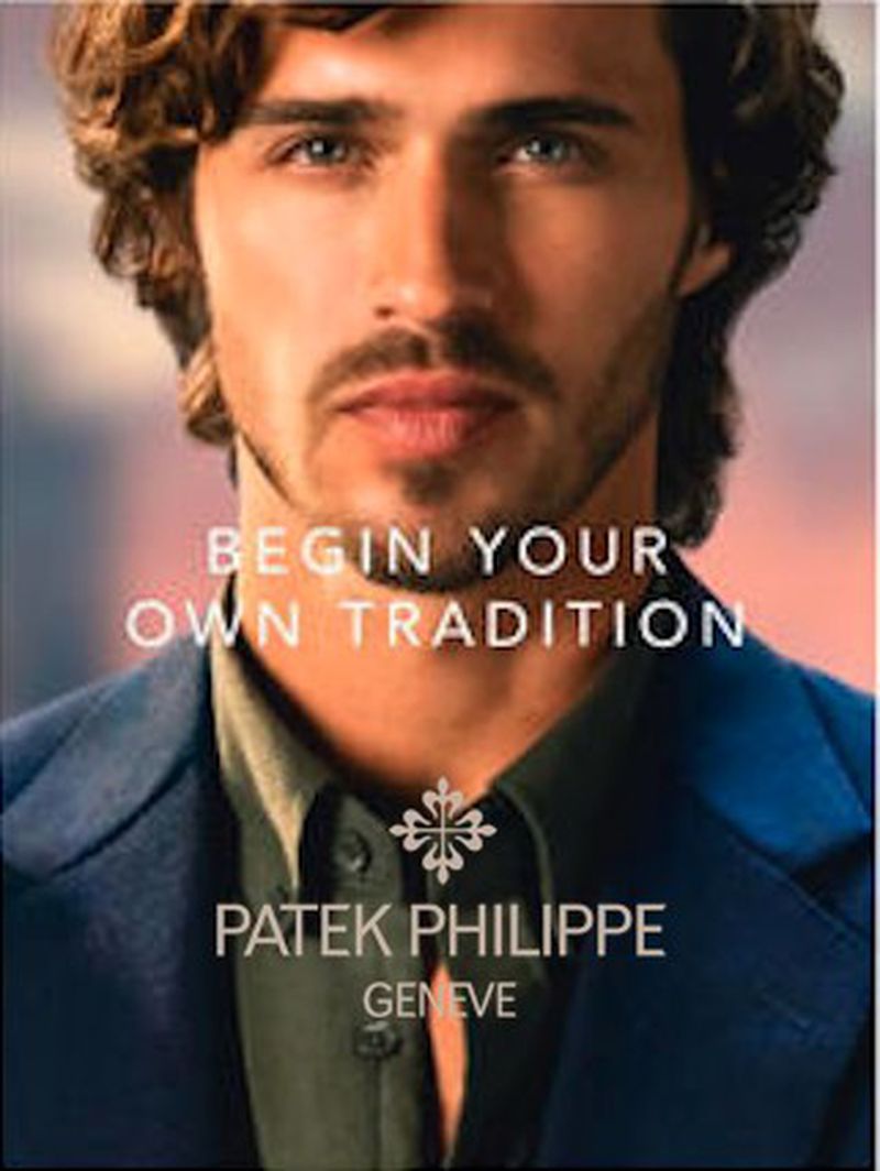 Close-up studio shot of an adult male model in a navy blazer and open-collared shirt, against a blurred background, promoting Patek Philippe.