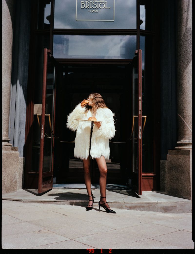 Woman in a white fur coat stands in front of The Bristol Hotel entrance, holding a plate with a croissant under natural lighting.