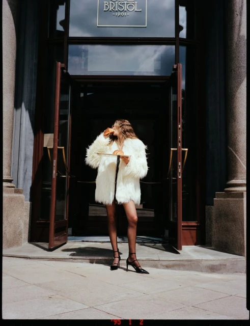 Woman in a white fur coat stands in front of The Bristol Hotel entrance, holding a plate with a croissant under natural lighting.