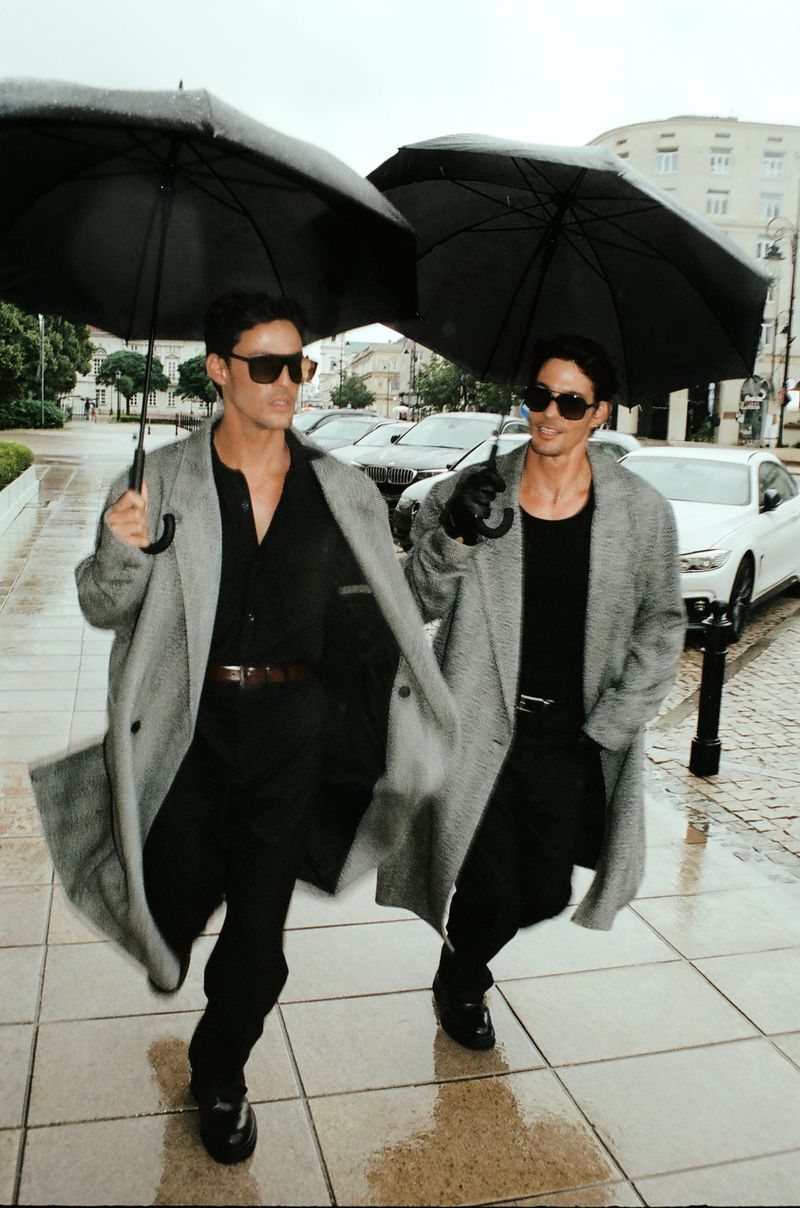 Two adult men in coats and sunglasses, walking in the rain with black umbrellas in a European city street. Fashionable, modern.