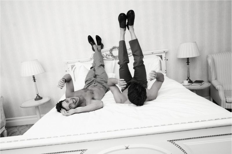 Two men lie upside down on a bed in an indoor room with a white bed and white walls and side tables, one man smiling and one facing away.