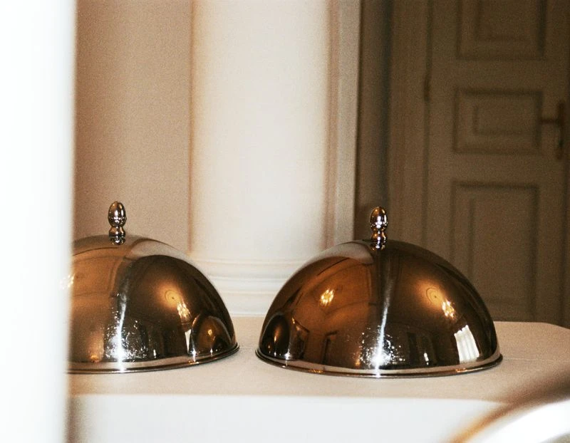 Two silver-domed food covers on a white tablecloth with a blurred background of a white column and a tan-colored door.