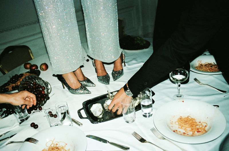 Two people standing on a formal dining table with plates of pasta, oysters, fruit, and drinks, wearing evening wear and high heels.