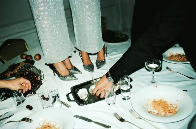 Two people standing on a formal dining table with plates of pasta, oysters, fruit, and drinks, wearing evening wear and high heels.