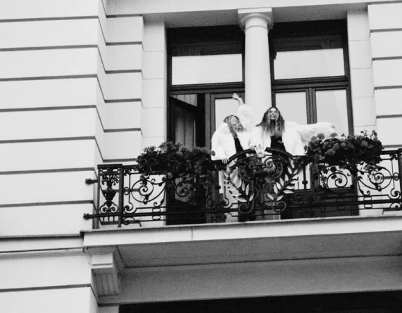 Two women in white outerwear pose on an ornate wrought-iron balcony with planters, against a European building with large windows.