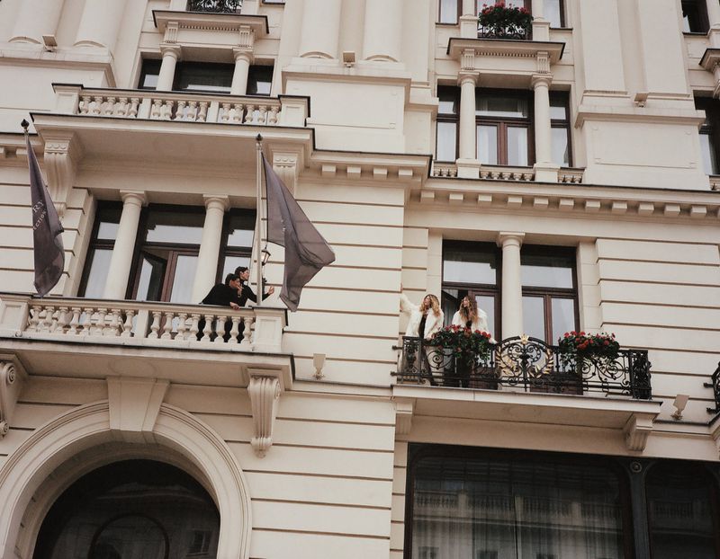 Two women waving from a building's balcony with flowers; two other figures on a nearby balcony; architectural detail and flags visible.