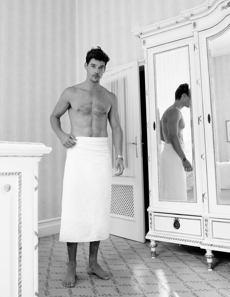 Man wearing a white towel in a light-filled room with antique white furniture, standing near a mirrored wardrobe, looking at camera.