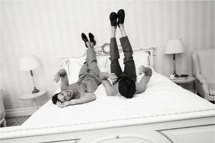 Two shirtless male models lying on a bed with legs elevated against a headboard for Kazar Studio A/W 24.