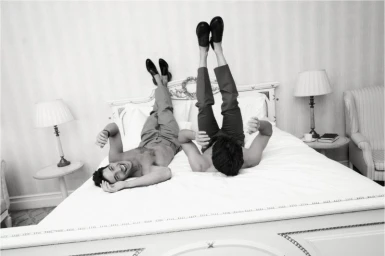 Two shirtless male models lying on a bed with legs elevated against a headboard for Kazar Studio A/W 24.