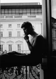 Shirtless man sitting on a balcony railing smoking a cigarette with a classical building in the background