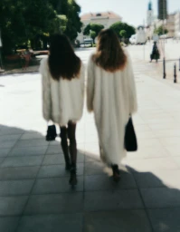 Two women with long hair walking away from camera wearing long white coats and holding black handbags on a sunny paved square