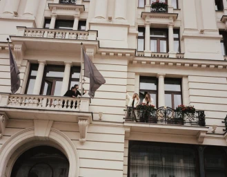 Models in outerwear standing on neoclassical building balconies with hanging flags and flower boxes.
