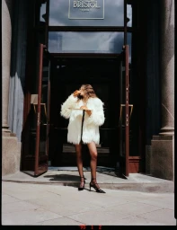 Model in fluffy white coat eating a croissant outside grand hotel entrance with dark double doors