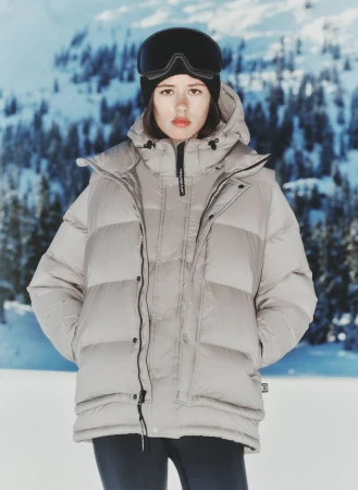 Woman in ski goggles and a puffer jacket stands against a snowy mountain backdrop on a bright day, creating a winter fashion editorial.