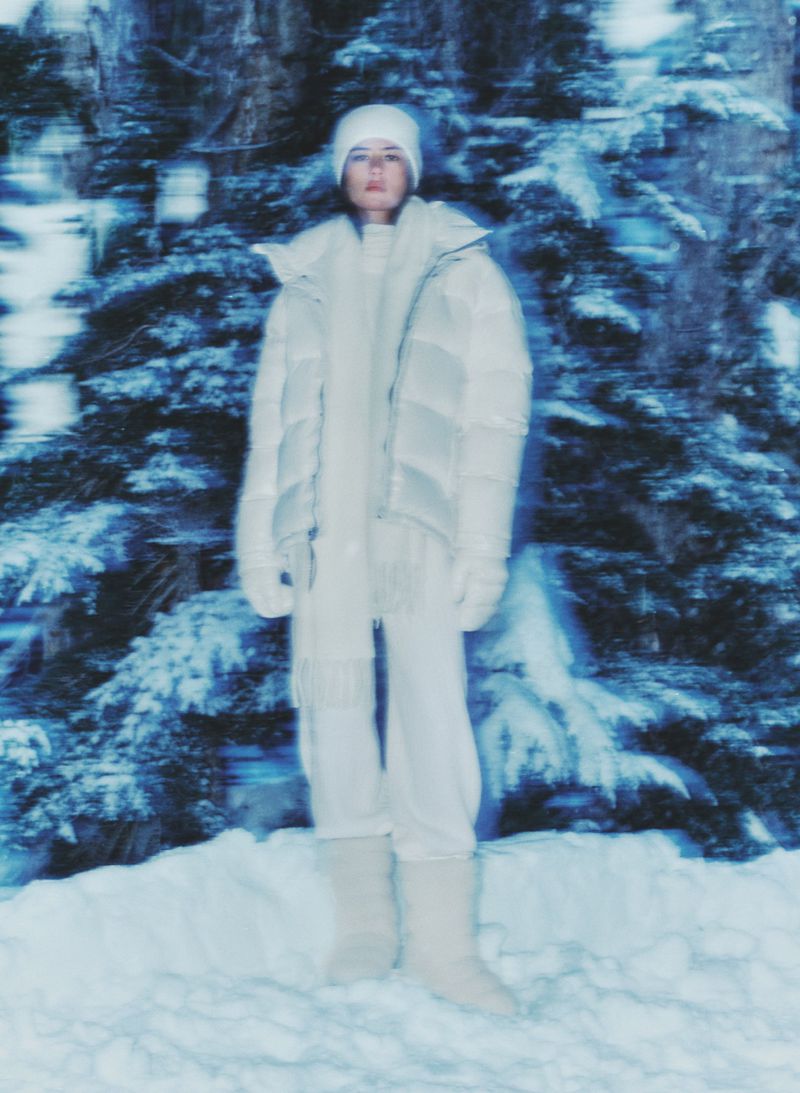 Woman in a puffy coat, knit hat, and boots stands in a snowy forest. Her white monochrome outfit contrasts with the wintry setting.