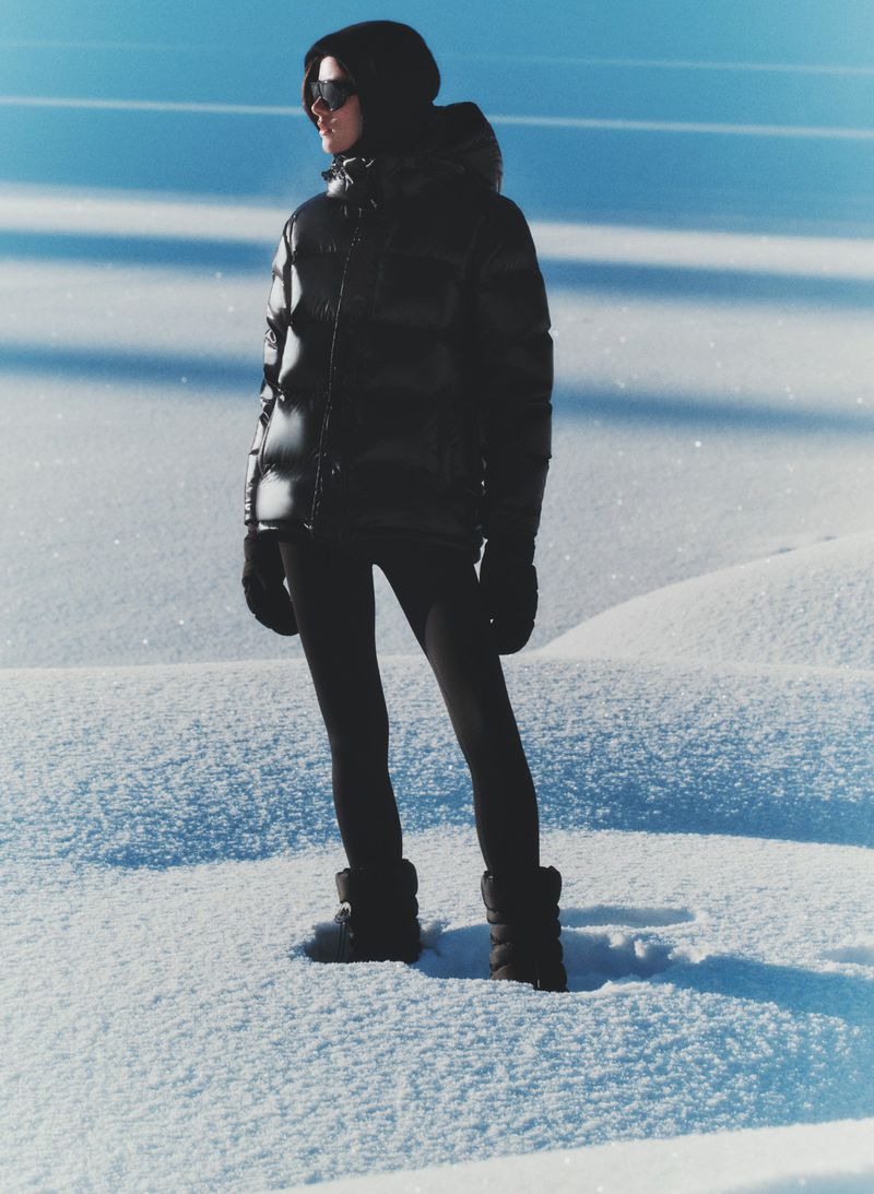 Woman in a black puffer jacket, leggings, and boots stands in a snowy field under a blue sky, creating a cool winter fashion image.