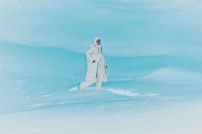 Woman in white winter coat walking in snow against light blue background. Full shot captures a winter fashion scene in a snowy environment.
