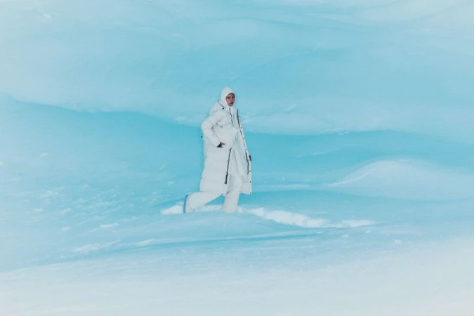 Woman in white winter coat walking in snow against light blue background. Full shot captures a winter fashion scene in a snowy environment.