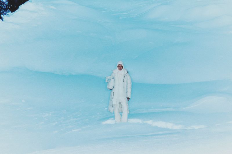 A lone woman in a white snowsuit stands in a vast, snow-covered landscape under soft lighting, holding a white object, creating a serene, wintery scene.