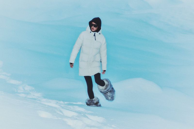 Woman in a white coat and fur boots walks in the snow. She wears dark sunglasses and a hat against a blue and white snowy backdrop.