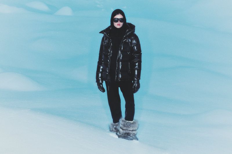 Person in black puffer jacket, balaclava and fur boots stands in snowy, light blue environment, looking at camera in full shot.