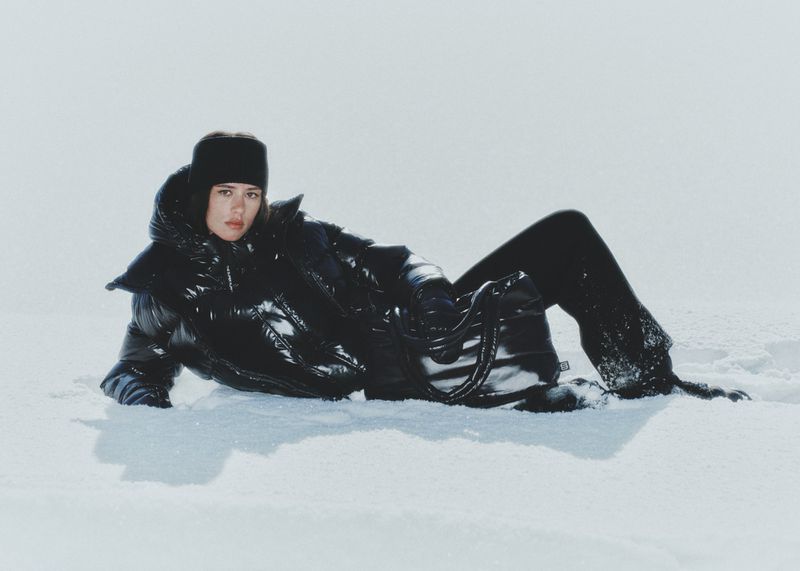 A person in a black puffer coat and matching pants lays in the snow against a light background in a fashion or editorial setting.