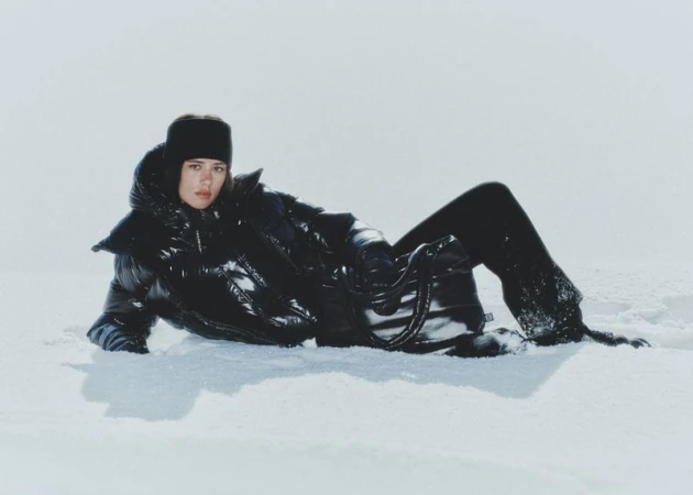 A person in a black puffer coat and matching pants lays in the snow against a light background in a fashion or editorial setting.