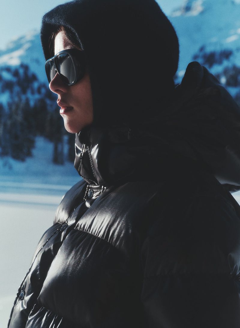 Adult in black puffer coat and sunglasses stands against a snowy mountain backdrop on a sunny day in outdoor setting.