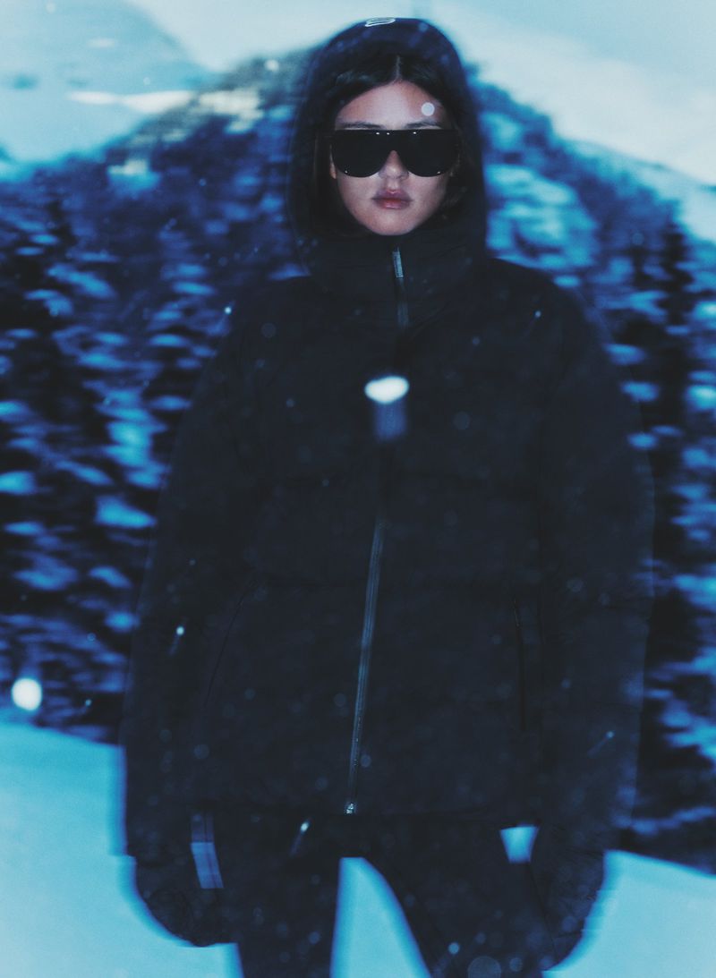 A woman in a black puffer jacket and sunglasses stands in a snowy landscape with blurred evergreen trees and a cool blue color grade.