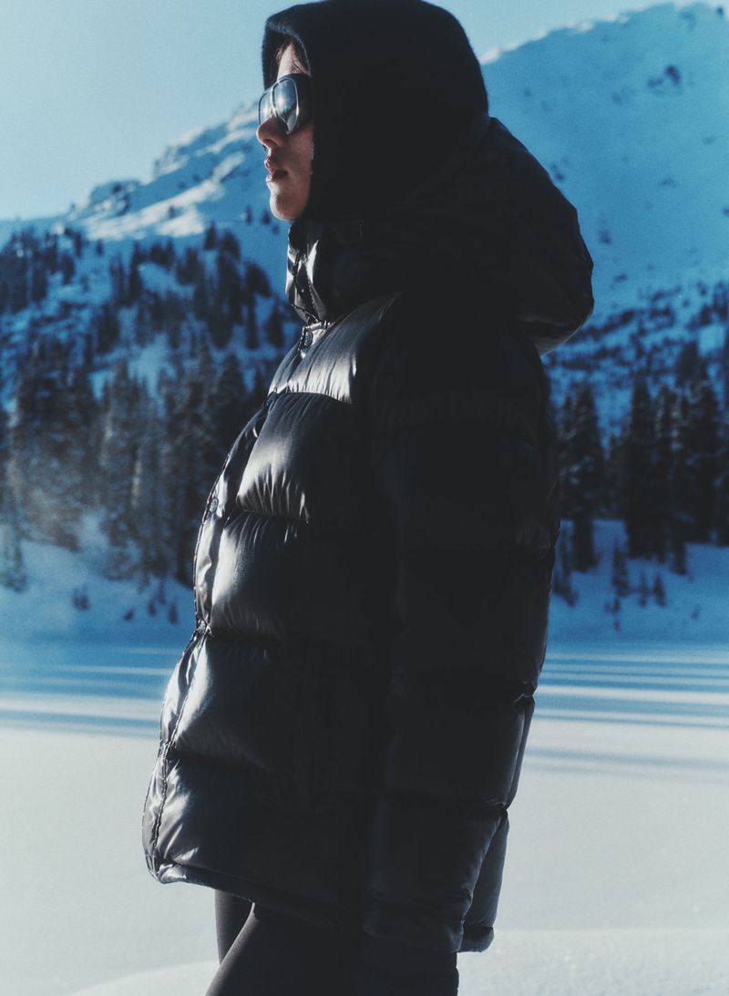 Side profile of a woman in a black puffer jacket and sunglasses standing outdoors in a snowy, mountainous landscape under a bright sky.