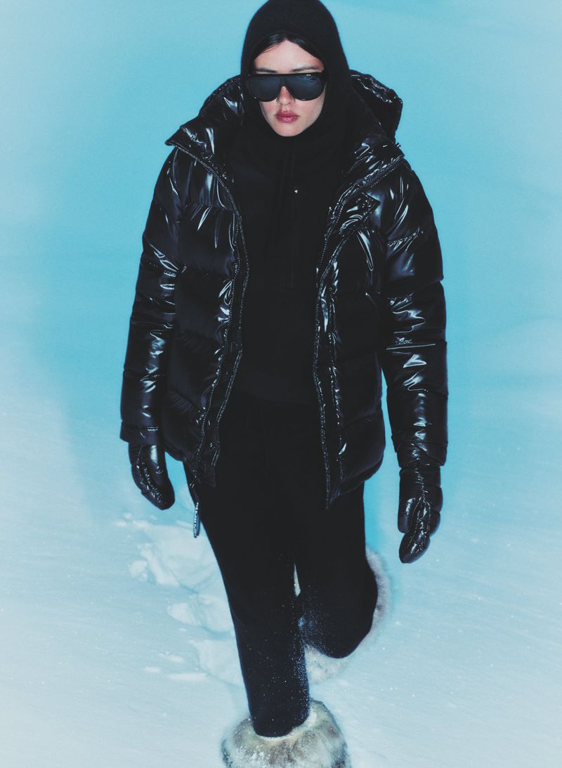 A woman in a black puffer jacket, leggings, gloves, and sunglasses stands in a snowy environment against a light blue backdrop.