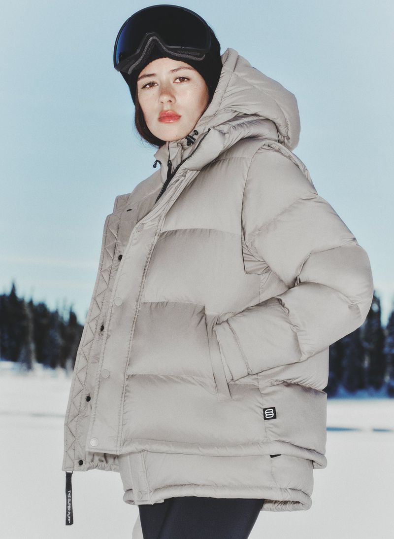 Woman in puffy grey jacket and snow goggles stands outside on a sunny day in winter.