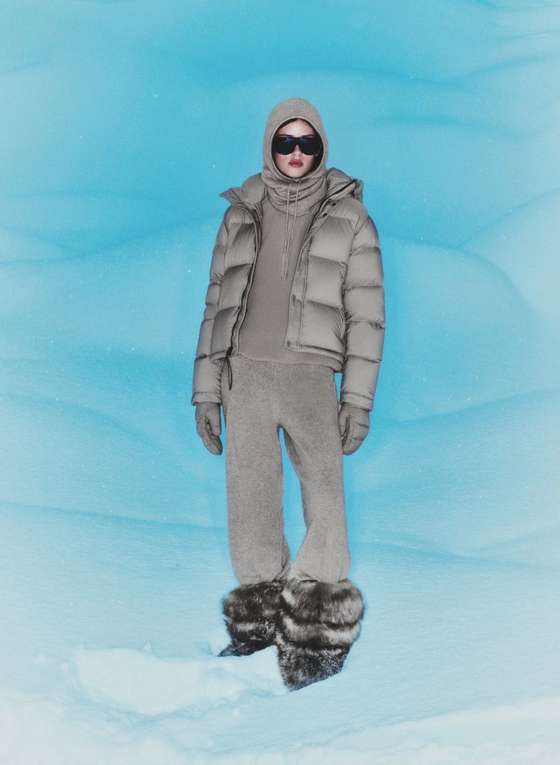 A person in a hooded puffer jacket and fuzzy boots stands in a snowy landscape against a blue backdrop, creating a winter fashion editorial.